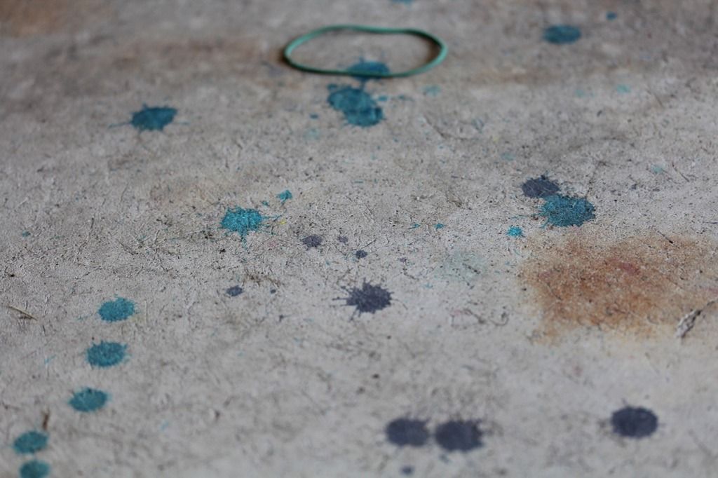 a concrete floor with drops of blue and turquoise dye on it, plus a green rubber band and a rust stain